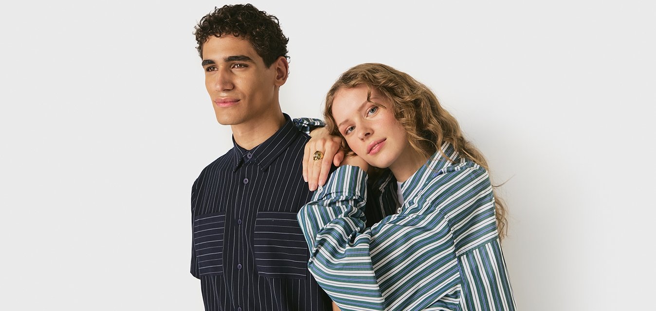 Muž a žena stojí vedle sebe, oba oblečeni v pruhovaných košilích; žena si položí hlavu a ruku na mužovo rameno na bílém pozadí.