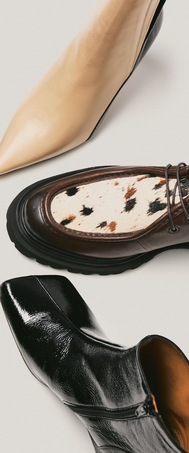 Tres pares de zapatos: un tacón puntiagudo de cuero beige, un zapato marrón con un panel textil con manchas, y una bota negra brillante hasta el tobillo con cremallera.