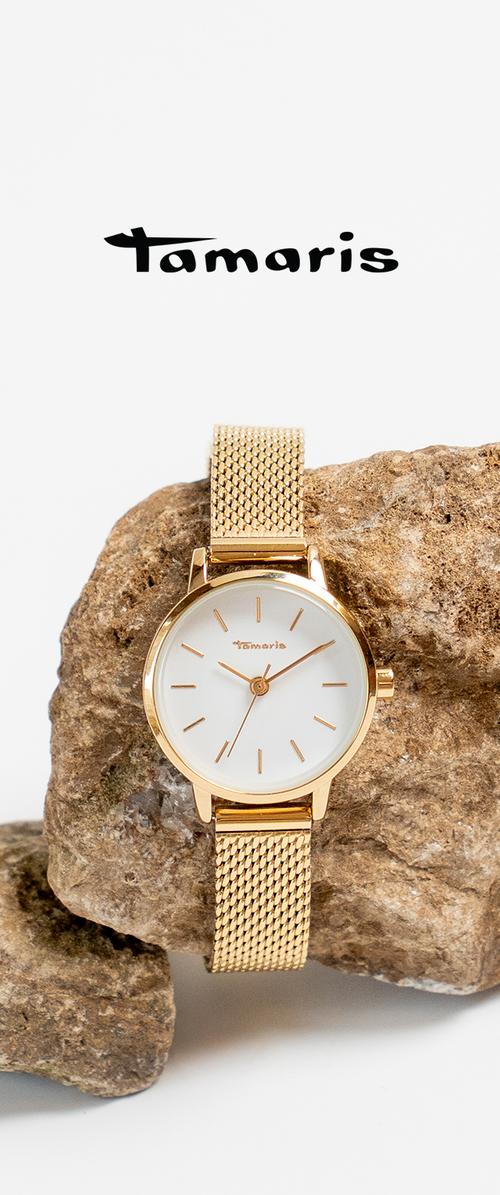 Reloj de pulsera dorado Tamaris con esfera blanca y correa de malla, colocado en posición vertical sobre un fondo de piedra rugosa, debajo del logotipo de la marca.