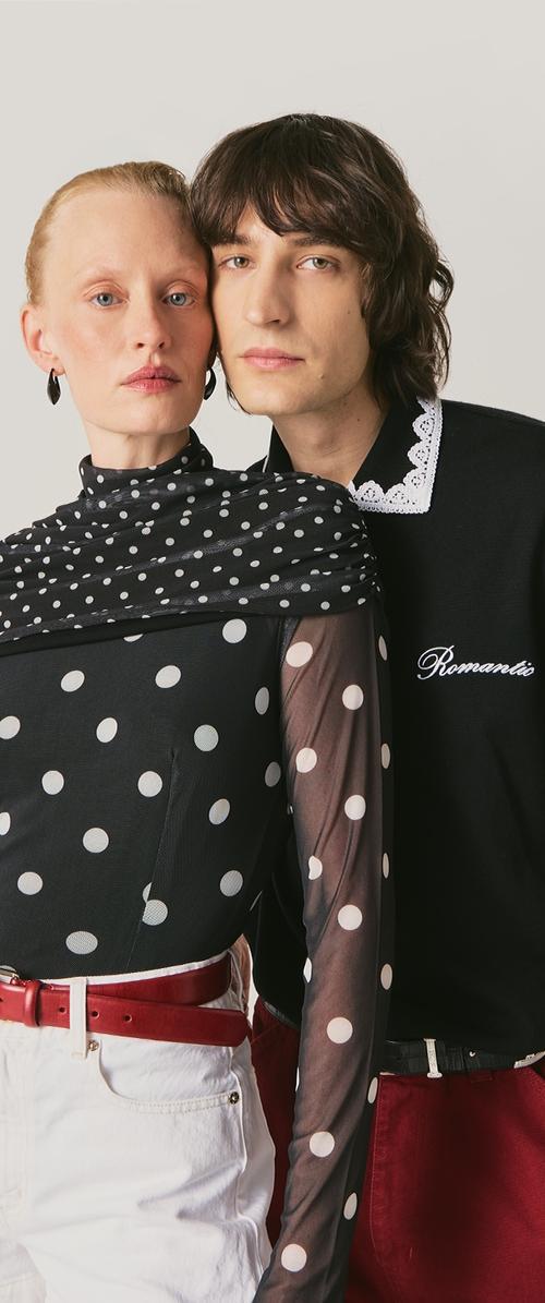 Zwei Personen dicht beieinander, eine trägt ein schwarzes, durchscheinendes Oberteil mit weißen Punkten, weiße Hose und roten Gürtel, die andere einen schwarzen Pullover mit weißem Spitzenkragen.