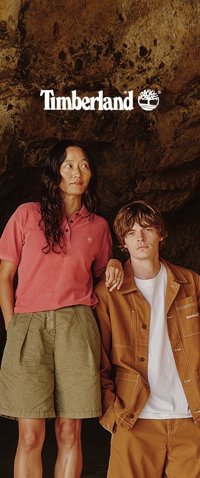 Mujer con polo rosa y pantalones cortos oliva está junto a un hombre con chaqueta y pantalones color marrón claro sobre un fondo rocoso con el logo de Timberland arriba.