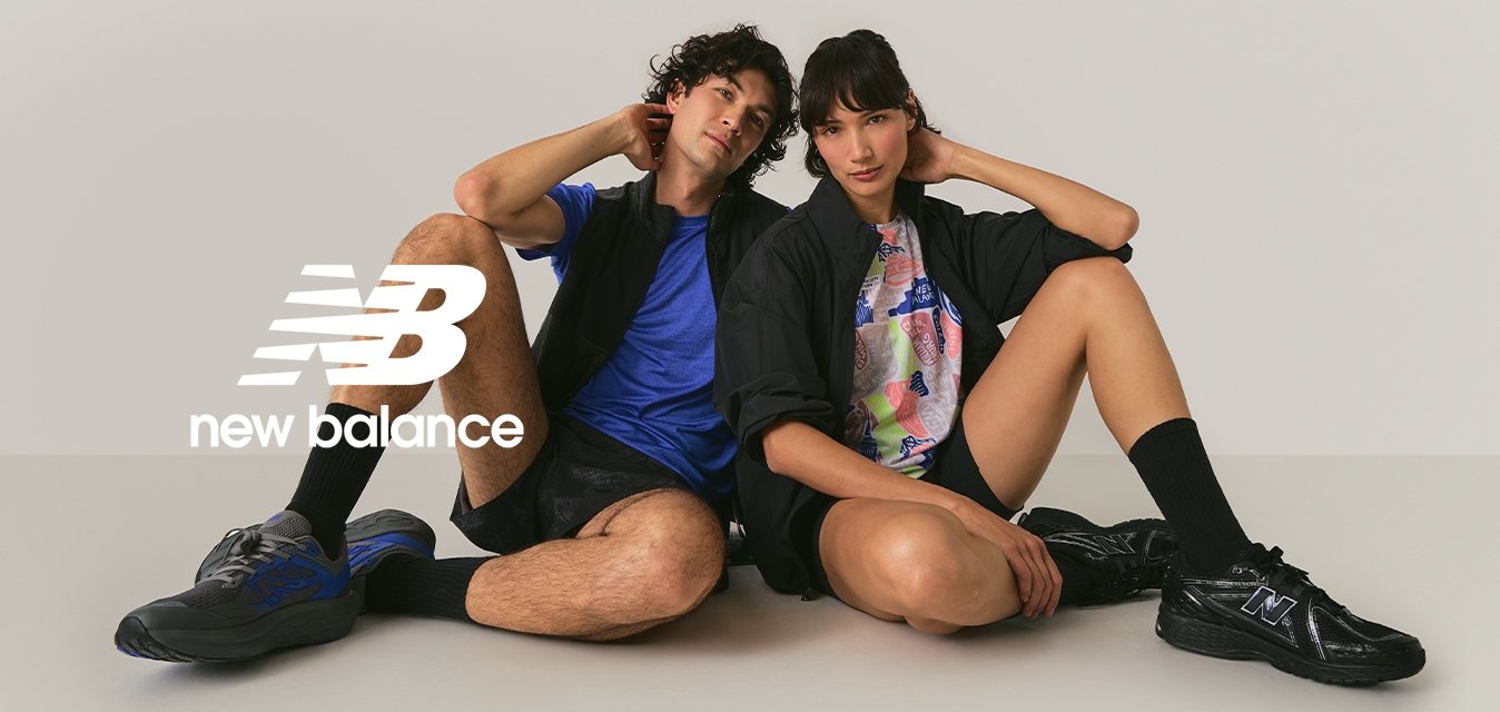 Dva mladí dospělí sedí na podlaze se skrčenýma nohama, oblečeni ve sportovním volném oblečení a teniskách, na pozadí jemně světle zbarvené stěny.