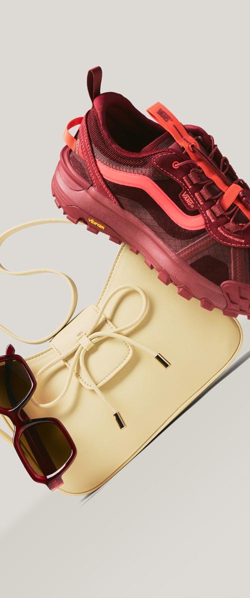 Zapatilla Vans marrón granate y naranja, bolso bandolera crema con detalle de lazo, y gafas de sol con montura marrón granate y lentes marrones sobre fondo claro.