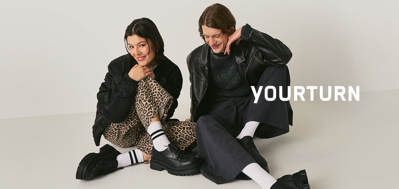 Dva lidé sedí na podlaze, mají na sobě černé bundy, bílé ponožky a robustní černé boty. Jeden má na sobě leopardí vzorované kalhoty, druhý široké tmavé kalhoty.