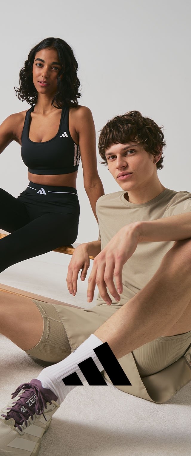 Mladá žena v černém sportovním oblečení a mladý muž v béžových šortkách a tričku sedí na zemi a na lavičce, pózují před jednobarevným pozadím.