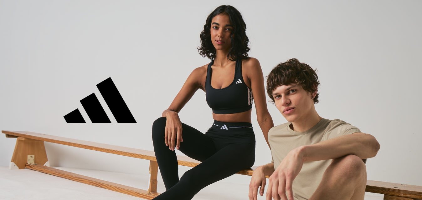 Dva mladí dospělí v sportovním oblečení sedí u dřevěných laviček v minimalistickém interiéru, oba mají na nohou tenisky Adidas.