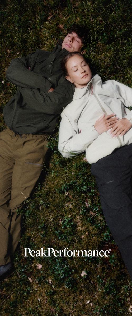 Zwei Personen liegen auf Gras mit geschlossenen Augen, tragen Outdoor-Jacken und Hosen, entspannt in einer natürlichen Umgebung.