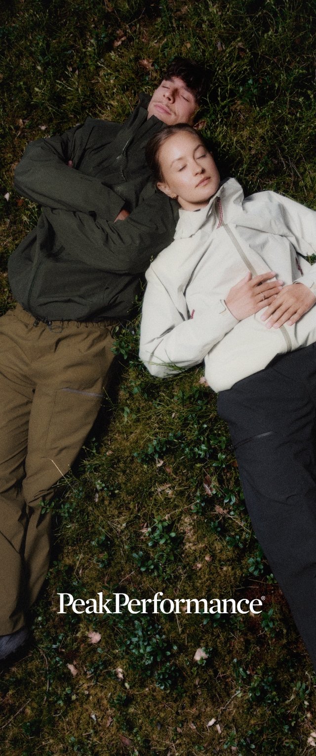 Dos personas tumbadas sobre la hierba con los ojos cerrados, vistiendo chaquetas y pantalones de exterior, relajadas en un entorno natural.