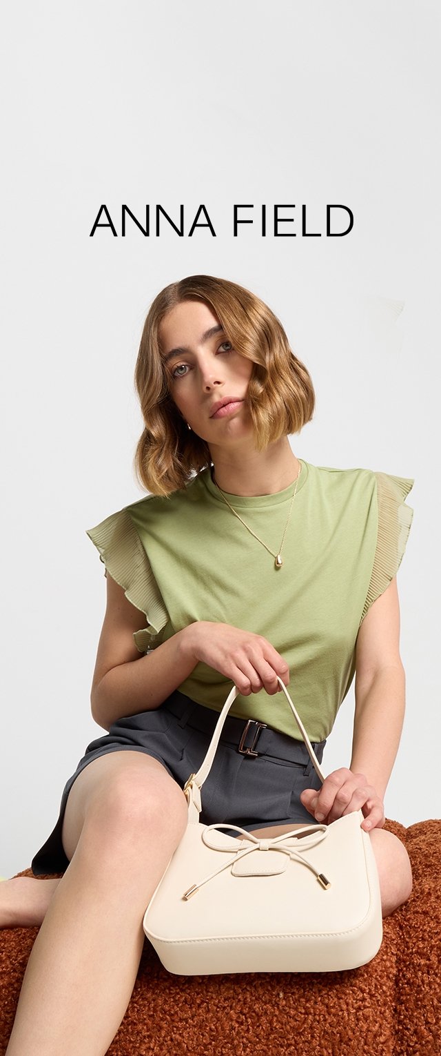 Hellgrünes Top mit Rüschendetail an den Schultern, kombiniert mit dunklen Shorts. Hält eine cremefarbene Handtasche mit einer Schleifenakzent und goldfarbener Hardware.