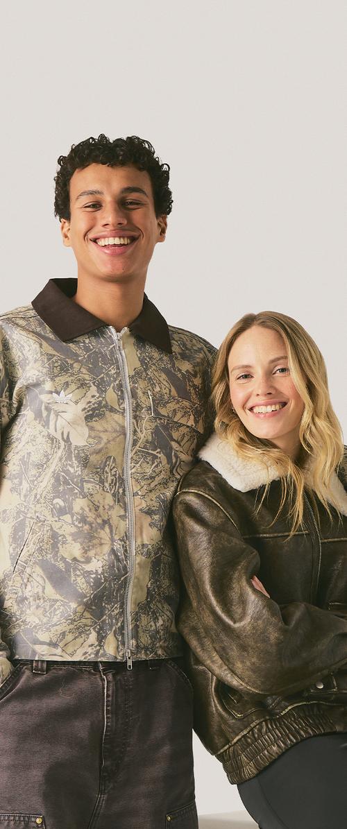 Giovane uomo e donna sorridenti in piedi vicini, indossano giacche casual con motivi e colletto in pelliccia, su uno sfondo chiaro uniforme.