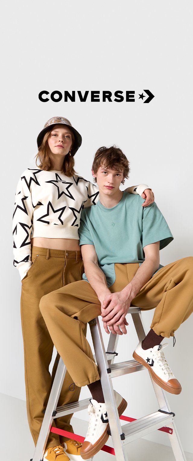 Weißer Sweatshirt mit schwarzem Sternmuster, hellgrünes T-Shirt, beigefarbene Hosen und beigefarbene Sneaker mit braunen Akzenten, alles auf einer Leiter.