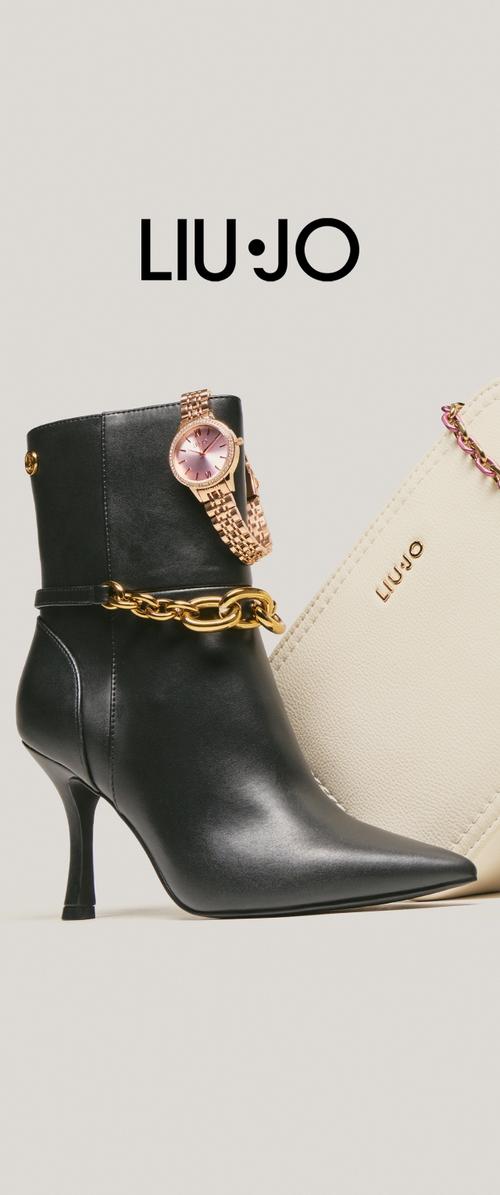 Botín negro de cuero con punta puntiaguda y tacón stiletto, decorado con un detalle de cadena dorada y combinado con un bolso crema. Marca LIU·JO visible.