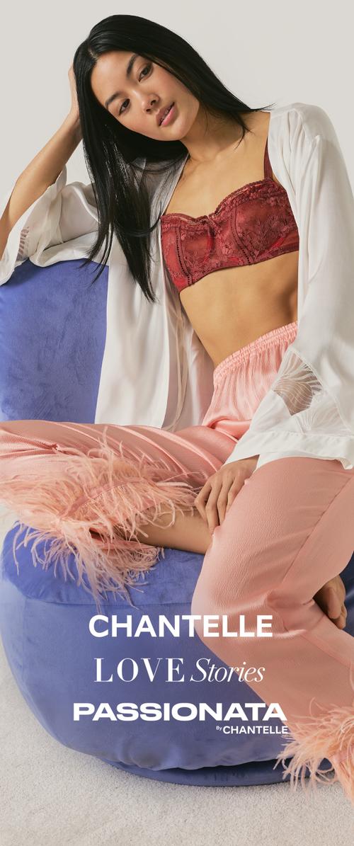 Frau mit langen schwarzen Haaren, trägt einen roten Spitzen-BH, eine weiße offene Strickjacke und rosa Federhosen, sitzt auf einem blauen Stuhl vor einem schlichten Hintergrund.