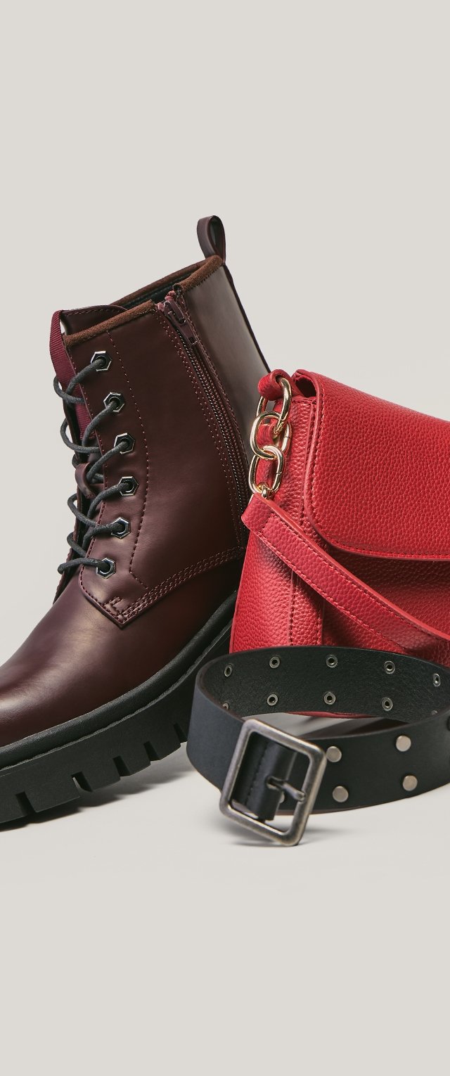 Stivaletti alla caviglia in pelle marrone con suole in gomma nere, abbinati a una borsa rossa con texture e a una cintura nera con borchie argentate e fibbia metallica.