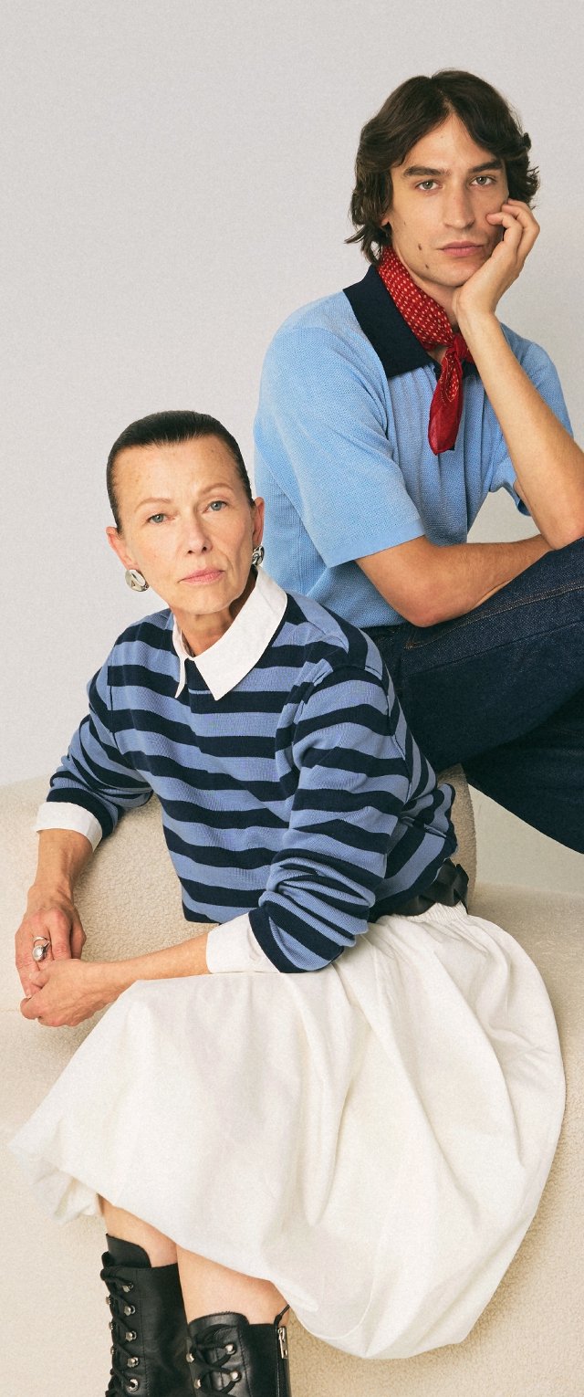 Femme âgée en pull rayé et jupe blanche assise, jeune homme en chemise bleue et écharpe rouge assis derrière elle sur une chaise beige sur fond neutre.