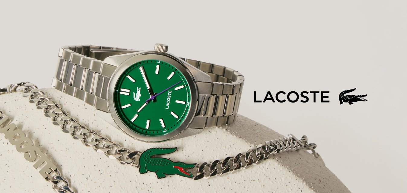 Stříbrné náramkové hodinky se zeleným ciferníkem a stříbrný náhrdelník s řetízkem, zdobený zeleným krokodýlím emblémem, na béžovém texturovaném povrchu.