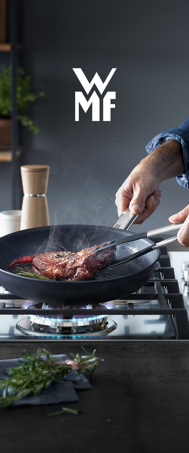 Schwarze Bratpfanne auf einem Gasherd mit einem Steak und Kräutern, Dampf steigt auf. Metallzange in einer Hand gehalten. Moderne Kücheneinrichtung mit einer hölzernen Pfeffermühle.