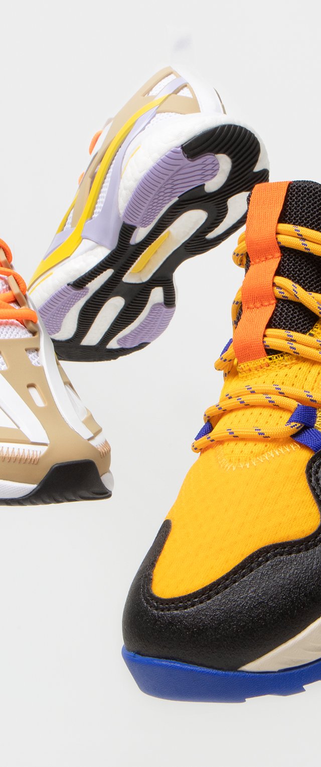 Bunt gefächerte Sportschuhe mit einem atmungsaktiven gelben Mesh-Obermaterial, schwarzen Akzenten, Gummisohle und orangefarbenen Schnürsenkeln sowie einem gemusterten Design.