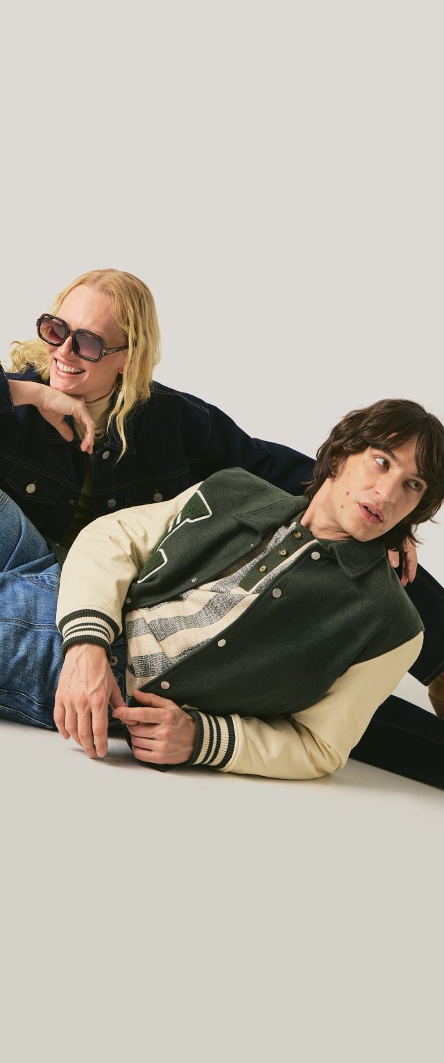 Deux jeunes adultes en vestes décontractées sont assis par terre ; l'un porte des lunettes de soleil et sourit, l'autre regarde sur le côté avec une expression neutre.