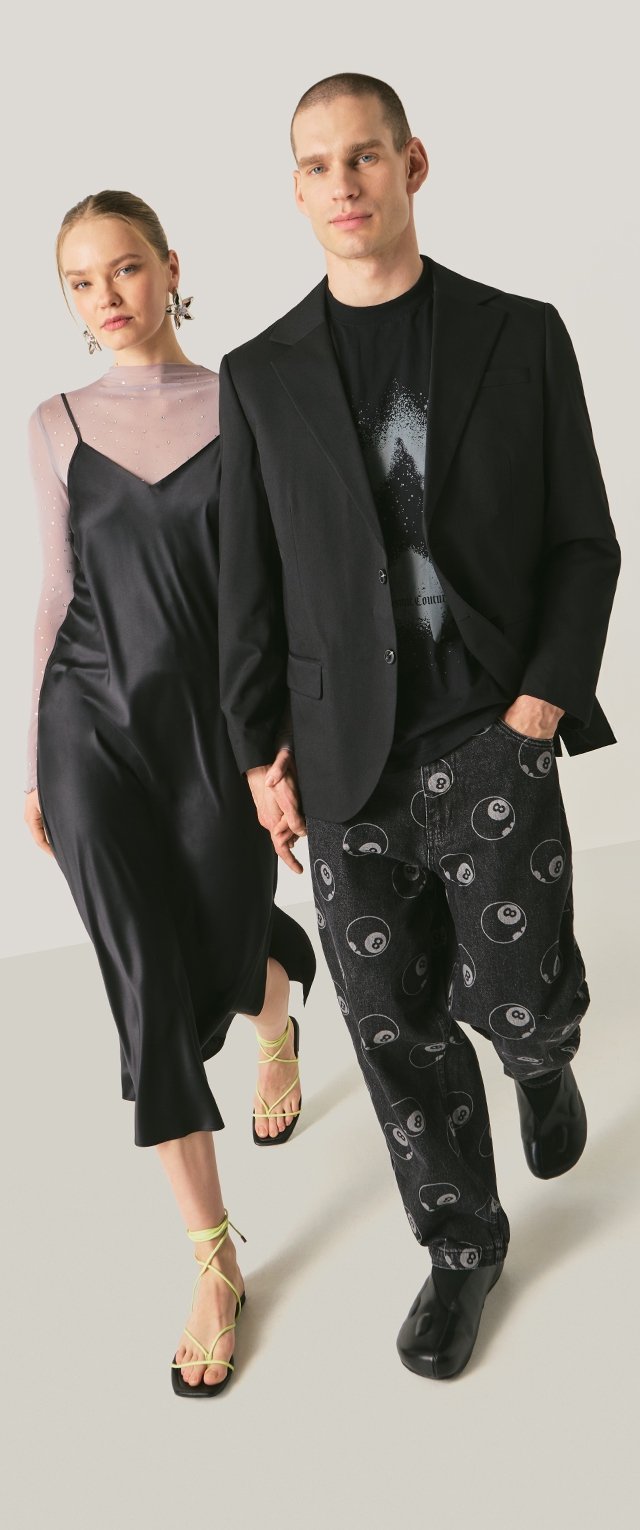 Kobieta w czarnej sukience na ramiączkach z przezroczystą długą bluzką na rękawach i neonowymi sandałami na cienkich paskach, mężczyzna w czarnym blezerze, koszulce z nadrukiem i czarnych spodniach z motywem biliardowej ósemki.