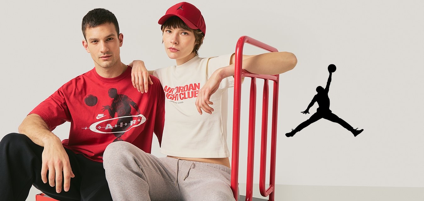 Dva mladí dospělí sedí na červeném plošinovém vozíku; jeden má na sobě červené tričko s grafikou, druhý bílé krátké tričko a červenou čepici.