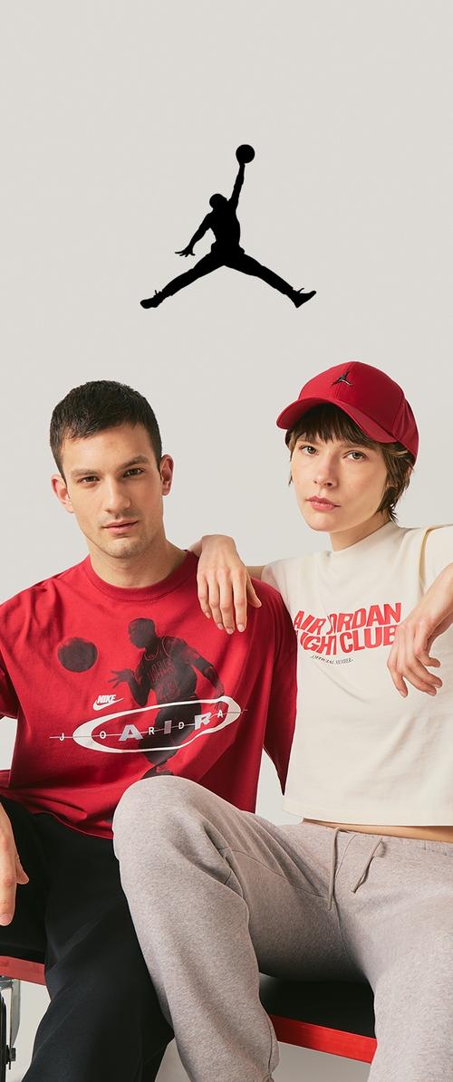 Dwóch młodych dorosłych w casualowej odzieży Jordan siedzi obok siebie, jeden z nich ma czerwoną czapkę, nad nimi czarne logo Jumpman na jednolitym tle.