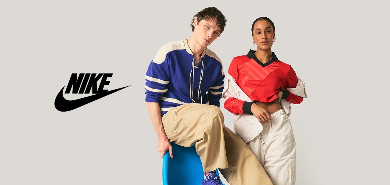 Dva mladí dospělí v neformálním oblečení pózují; jeden sedí na modré moderní židli, druhý stojí s rukou v kapse před jednoduchým pozadím.