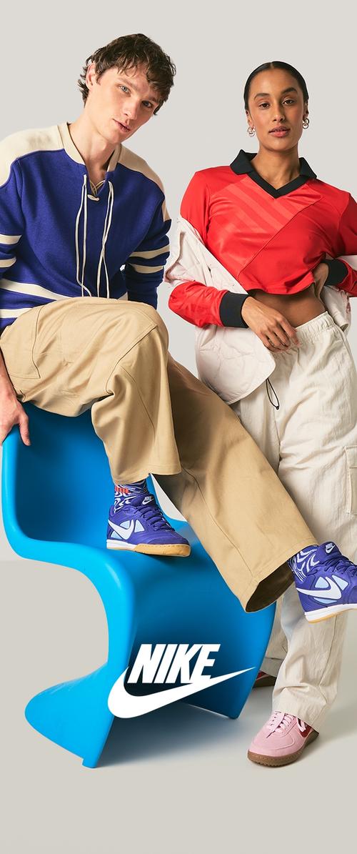 Mann in blauer und beiger Sportkleidung sitzt auf einem blauen Stuhl, Frau in rotem bauchfreiem Oberteil und beigen Hosen steht, beide tragen Nike-Schuhe.