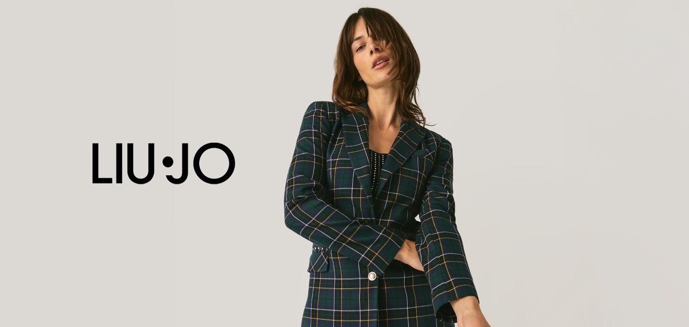 Žena pózující v kostkovaném saku a černých kalhotách s flitry na jednobarevném světlém pozadí.