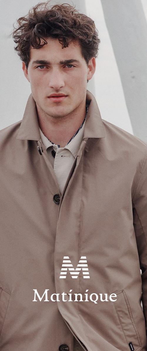 Junger Mann mit lockigem Haar, der einen beigen Trenchcoat über einem hellen Hemd trägt und vor einem neutralen, hellen Hintergrund steht.