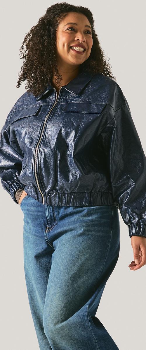 Leende kvinna med lockigt hår, iklädd en blank marinblå jacka med dragkedja och blå jeans, står med en hand i fickan.