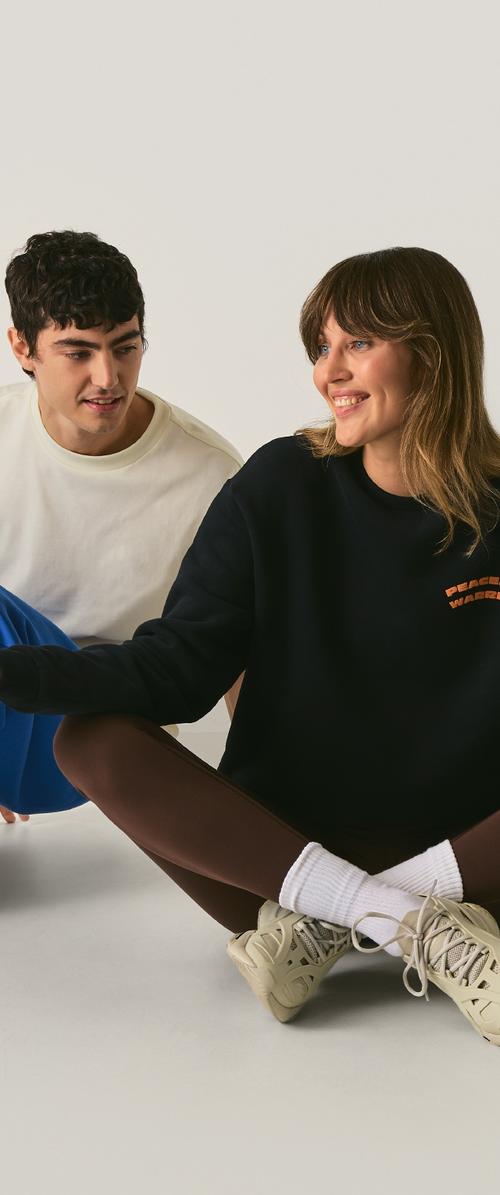 Joven vestido con sudadera blanca y pantalones azules sentado al lado de una mujer sonriente con sudadera negra, leggings marrones, calcetines blancos y zapatillas beige.