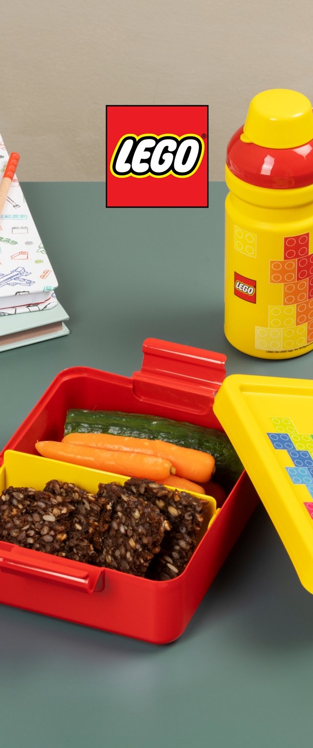 Scatola per il pranzo rossa LEGO con coperchio giallo, divisa in sezioni che contengono cetriolo, carota e snack quadrati. Include una bottiglia gialla coordinata.