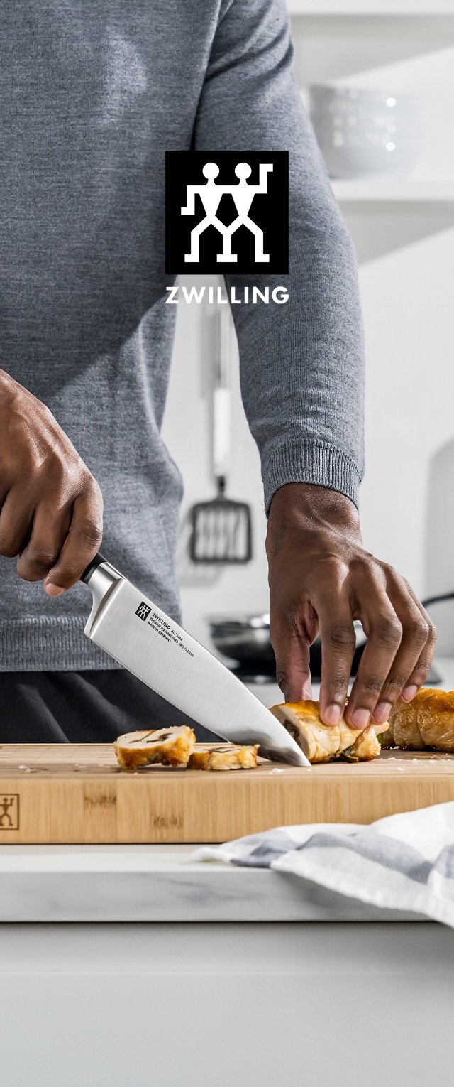 Couteau de chef ZWILLING avec lame en acier inoxydable coupant du poulet cuit sur une planche à découper en bois. Un pull gris et un tissu rayé sont visibles.
