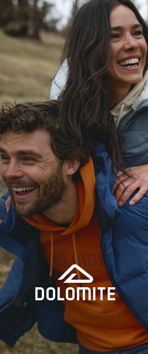 Un hombre sonriente con una sudadera naranja y una chaqueta azul lleva a una mujer riendo a sus espaldas al aire libre en un entorno natural.