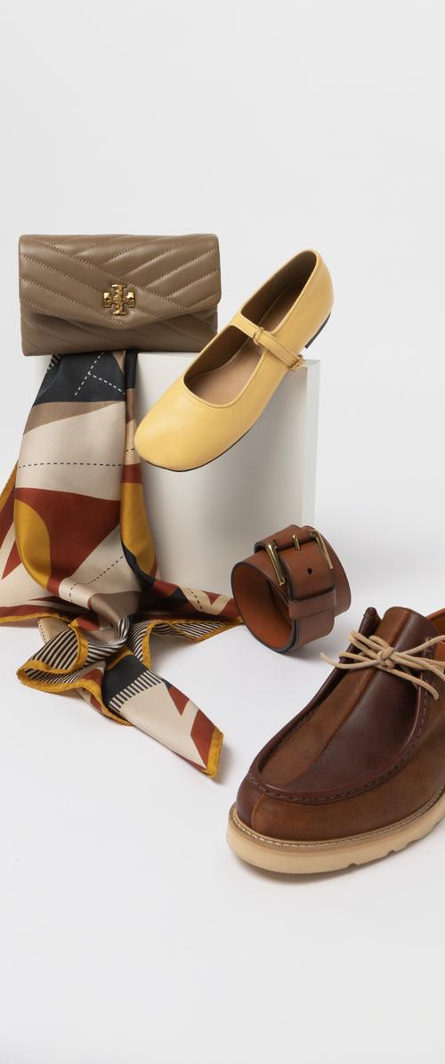Scarpe in pelle marrone con suola color crema, ballerine in pelle gialla, un portafoglio trapuntato beige, una sciarpa con motivo e una cintura in pelle marrone con dettagli dorati.