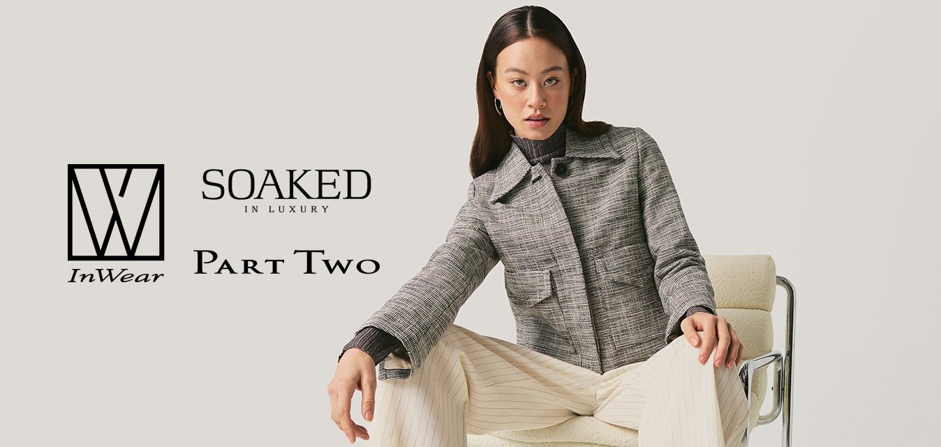 Žena s černými vlasy v šedé bundě a krémových kalhotách sedí na židli s kovovým rámem na jednobarevném bílém pozadí.