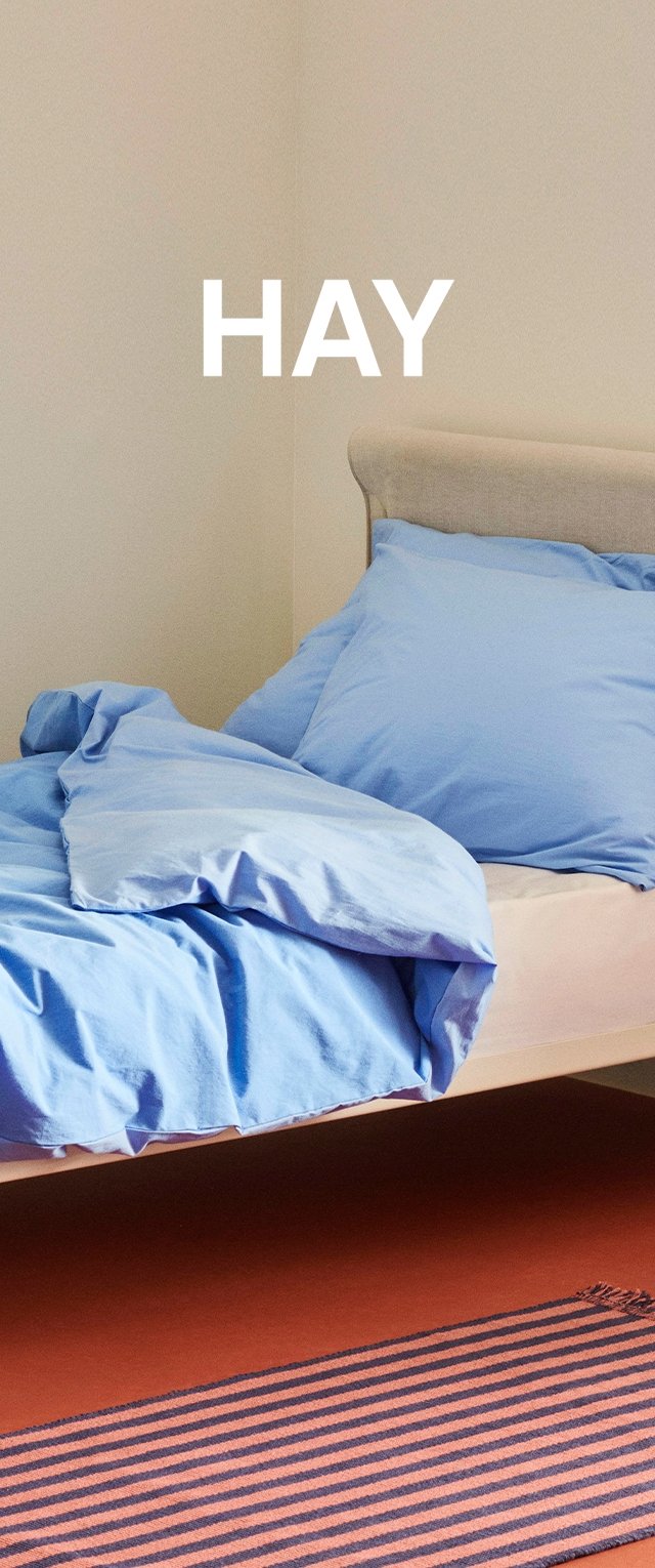 Himmelblaues Bettzeug liegt auf einem Bett mit einem beigen Kopfteil, das an einer hellen Wand steht. Ein gestreifter Teppich in Rost- und Marineblau ist sichtbar.