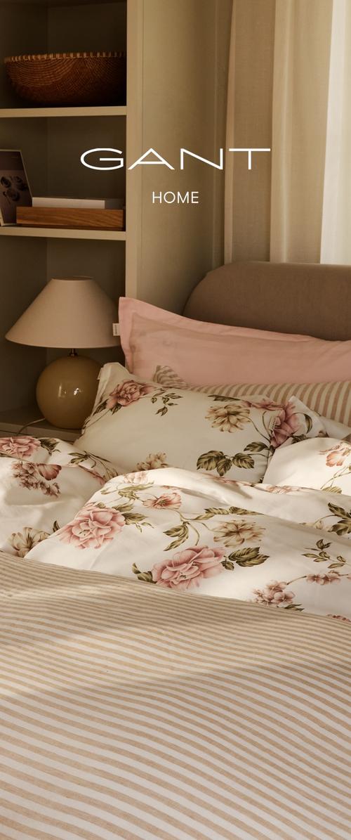 Cama con ropa de cama floral y de rayas, cabecero beige, lámpara beige sobre la mesita de noche y estantes con cesta y foto enmarcada con luz suave.