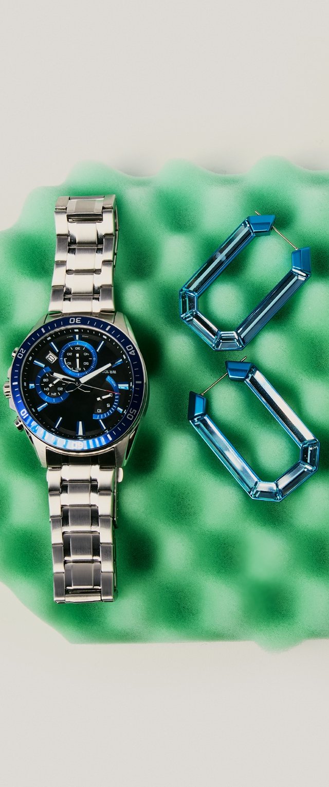 Strieborné kovové hodinky s modrými akcentmi a čiernym ciferníkom, spolu s dvoma priesvitnými modrými geometrickými náušnicami na zelenej pene.