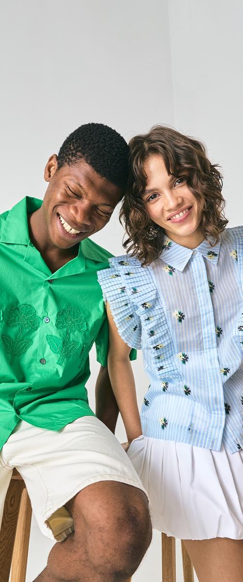Un homme portant une chemise verte brodée et un short blanc, à côté d'une femme vêtue d'un haut bleu à rayures avec des motifs floraux et des manches volantes.
