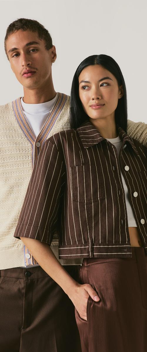 Deux personnes posent ensemble. L’homme porte un cardigan en maille crème et un pantalon marron. La femme porte une veste marron à rayures et un pantalon.