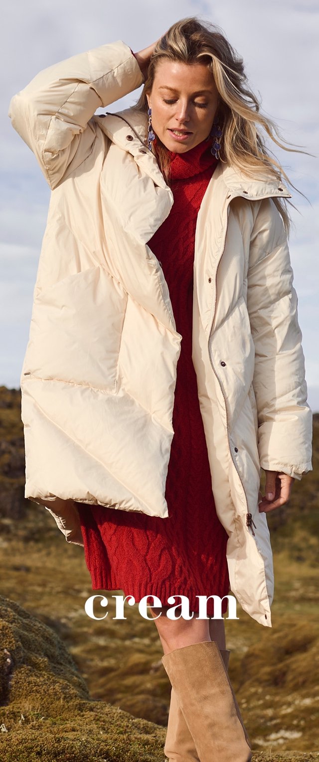 Vrouw in een crèmekleurige pufferjas, een rode gebreide jurk en tan suède laarzen, staande op een mosachtige ondergrond in de buitenlucht.