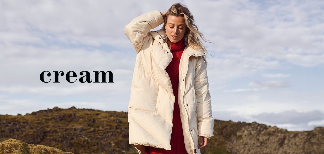 Nafoukaný bílý kabát s velkými kapsami, nošený přes žebrované červené šaty, doplněný o světlé vysoké boty, na pozadí mechového krajinného rázu.