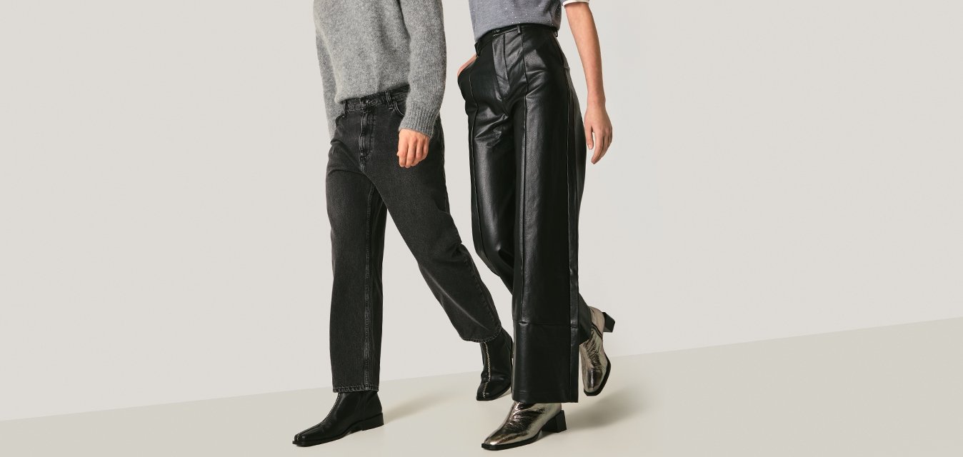 Muž a žena kráčí vedle sebe, mají na sobě šedá trička, černé kalhoty a kotníkové boty, na pozadí je jednoduché světlé pozadí.