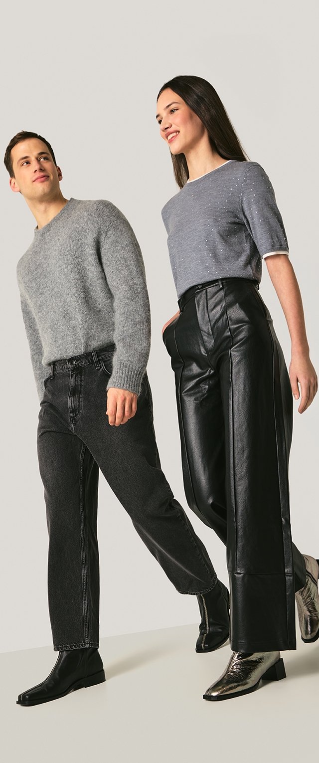 Pulover gri tricotat, pantaloni evazați din piele neagră și cizme metalice, cu finisaj texturat. Modelul pozează pe un fundal neutru.