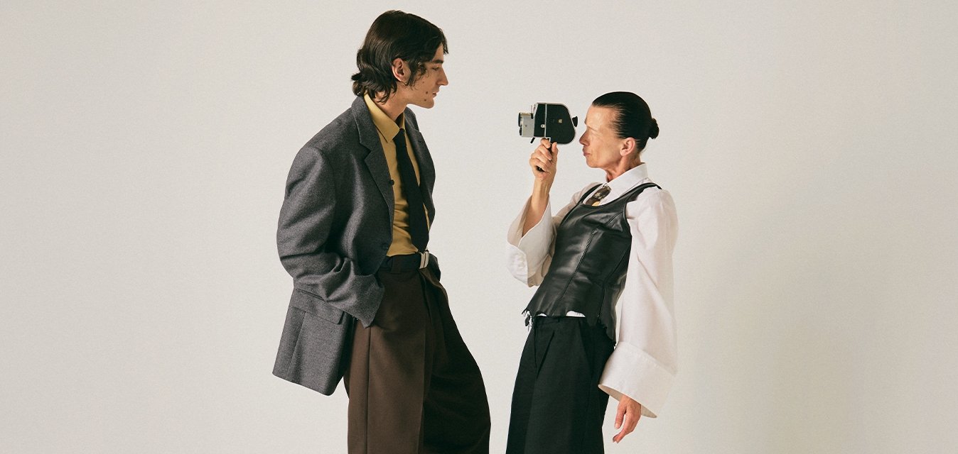 Dva lidé stojí na světlé podlaze proti jednoduché zdi; jeden drží vintage fotoaparát a míří jím na druhého, který pozorně sleduje.