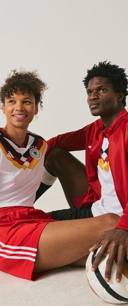 Deux joueurs de football en maillots d'Allemagne assis, l'un tenant un ballon de football, sur un fond neutre uni.