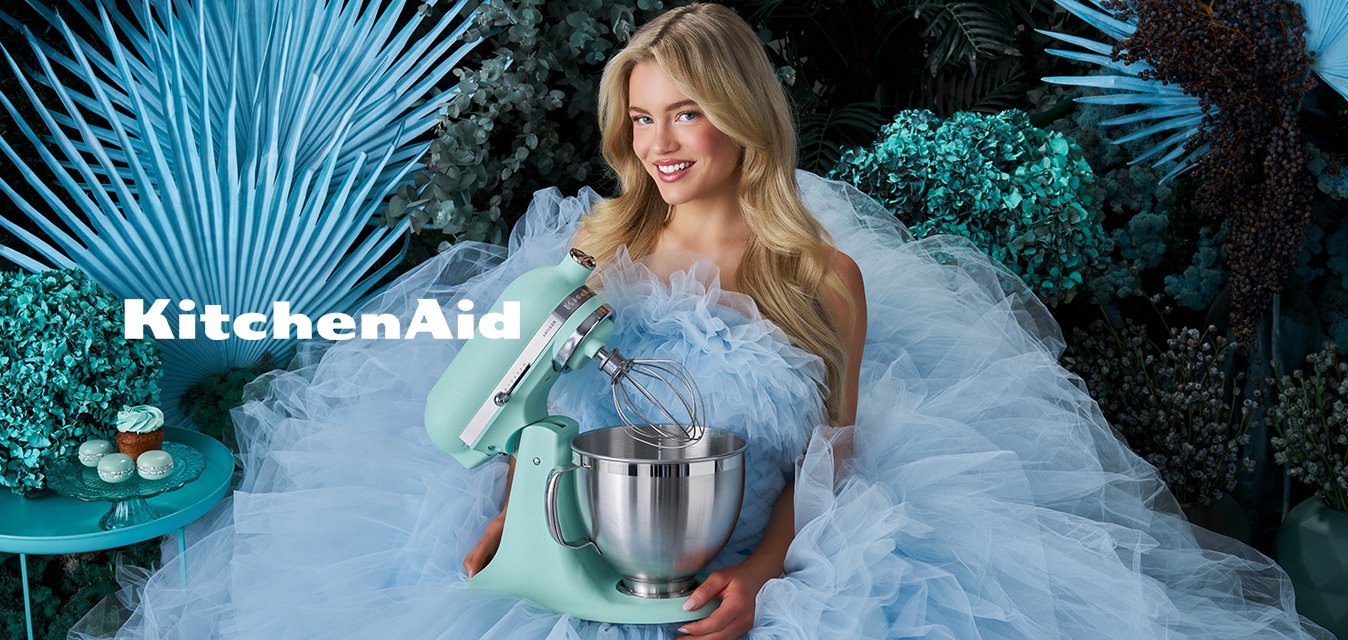 Žena v objemných světle modrých tylových šatech drží mintově zelený stolní mixér, obklopená modrozelenou listovou vegetací a cupcaky na malém stolku.