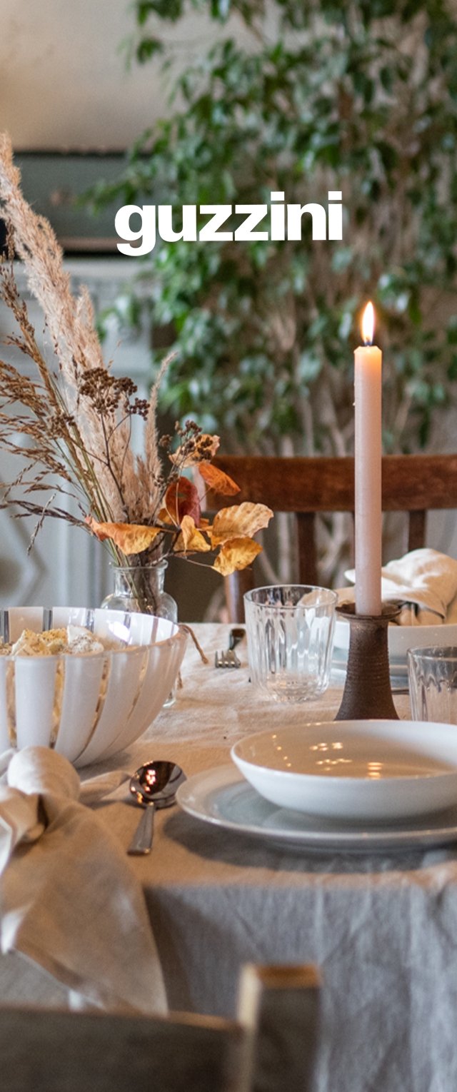 Matbord med vita tallrikar, glas, ett tänt ljus, torkad blomsterarrangemang i mitten och vikta servetter på en ljus duk.